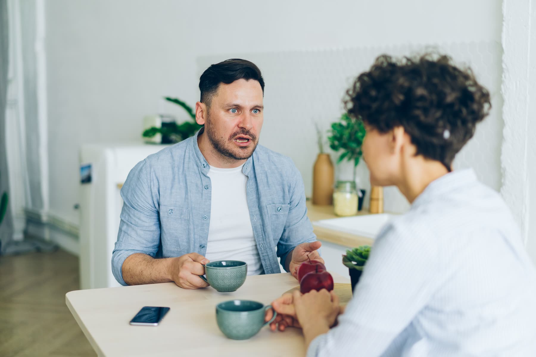 Two people at a kitchen table with coffee, mid-conversation, one listening while the other speaks.