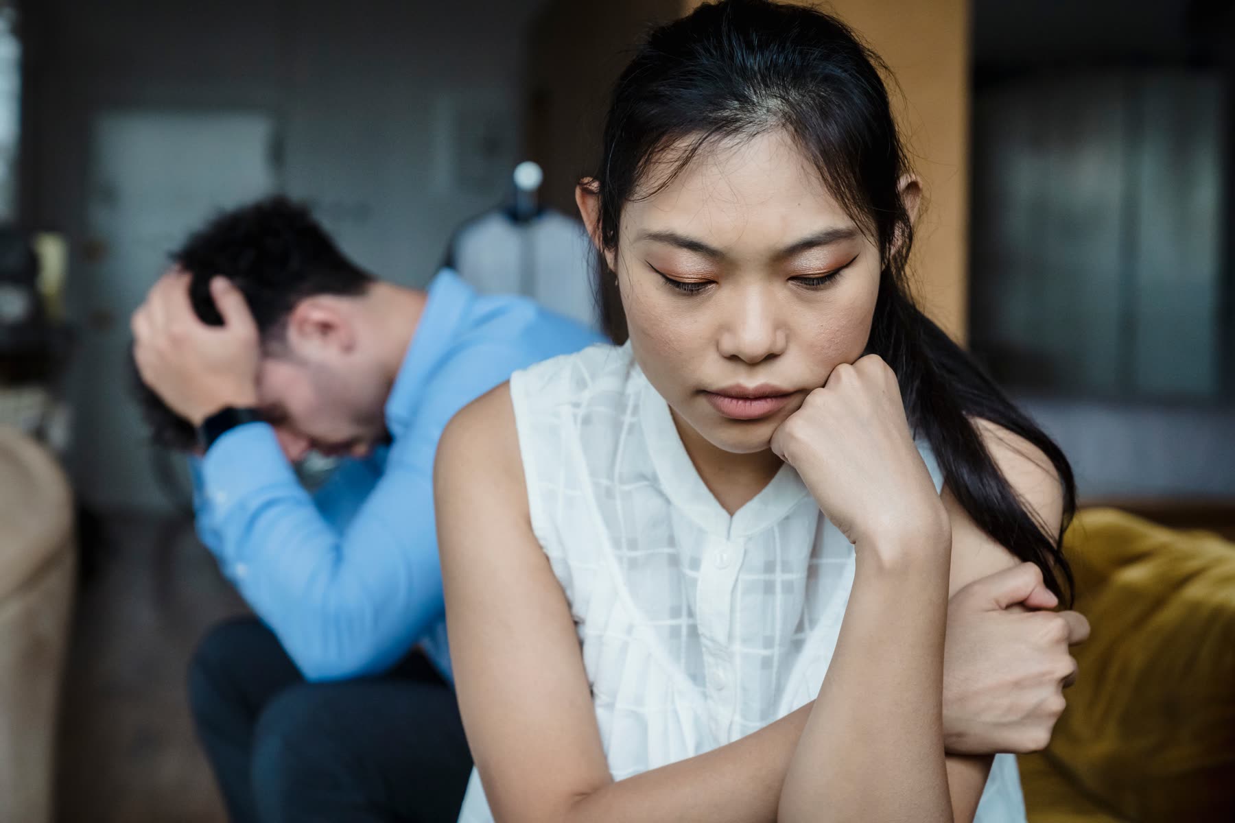 A couple sitting apart on a sofa after an argument, one with their head in their hands, neither looking at the other.